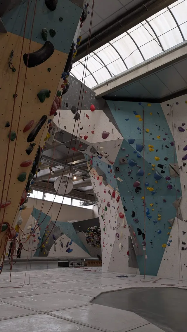 Salle d'escalade : grands murs de voies couverts de prises colorées, cordes en place, verrière au plafond